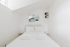 Quarto branco com uma cama, um banquinho pequeno, um espelho e uma estante com duas luminárias.