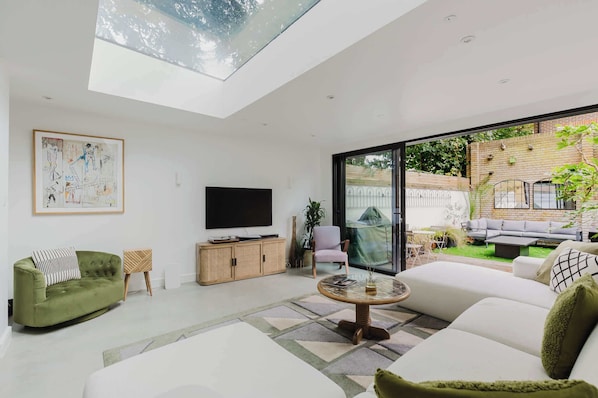 A modern living room with a large glass door leading to a garden, a flat-screen TV, a green sofa, and a white sectional sofa.
