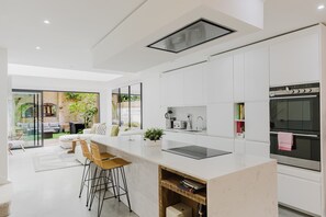 A modern kitchen with white cabinetry, a large island with stools, a built-in oven, and a view of a brick exterior.