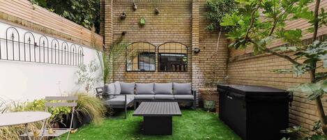 A backyard with a brick wall, a wooden deck, a grey sofa, a black table, and a hot tub.