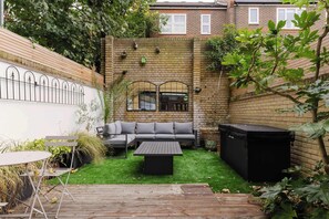 A backyard with a brick wall, a wooden deck, a grey sofa, a black table, and a hot tub.
