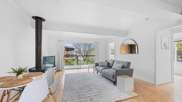 A modern living room with a sofa, a TV, a fireplace, and a view of a balcony with a wooden deck and greenery. - Serene 2BR Retreat in Goughs Bay Lake Eildon (Goughs Bay)