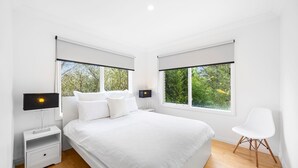 A modern bedroom with a large bed, two bedside tables, a nightstand, a chair, and two windows with blinds. - Serene 2BR Retreat in Goughs Bay Lake Eildon (Goughs Bay)