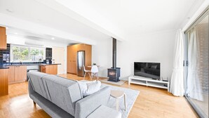 A modern living room with a grey sofa, a TV on a white stand, a black wood-burning stove, and a kitchen area with wooden cabinets and a window. - Serene 2BR Retreat in Goughs Bay Lake Eildon (Goughs Bay)
