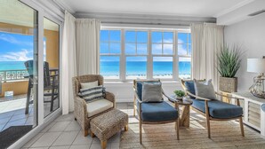 Amplia habitación con grandes ventanales con vistas a una playa, amueblada con sofá, sillón y reposapiés.