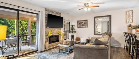 A spacious living room with a grey sofa, a coffee table, and a TV mounted on the wall. There is a balcony with outdoor furniture and a view of trees.