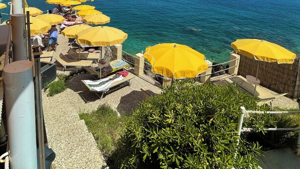 Une terrasse en bord de mer, agrémentée de parasols jaunes, de chaises longues et offrant une vue sur la mer et les montagnes lointaines.