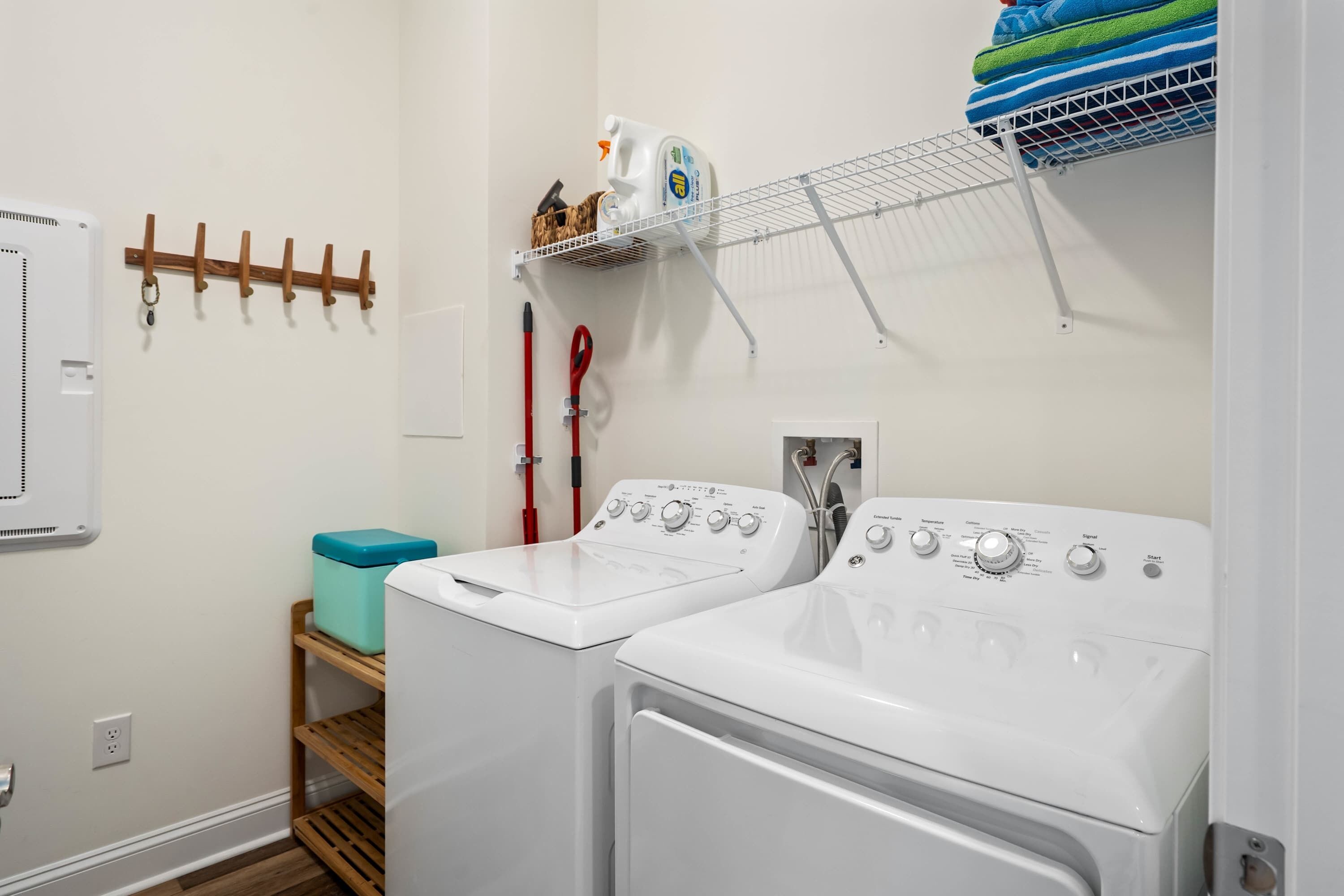 A laundry room with two front-loading washing machines, a drying rack, a small shelf with cleaning supplies, and a wooden stool.