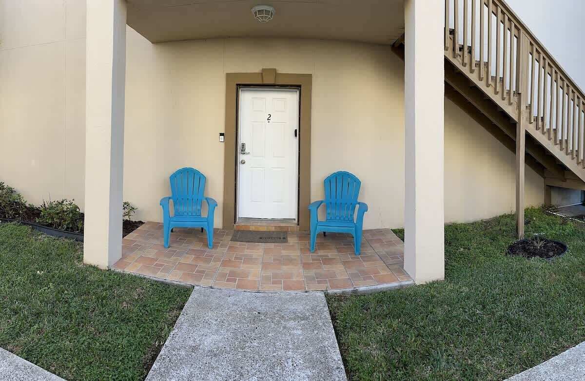 A building entrance with a white door marked '2', two blue chairs, a concrete pathway, and a staircase to the right.