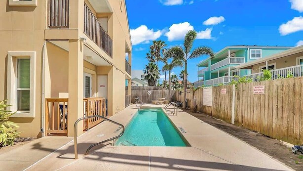 A pool area with a wooden fence, a clear blue sky, and a building with balconies. - Lovely Beach and Pool Condo (South Padre Island)