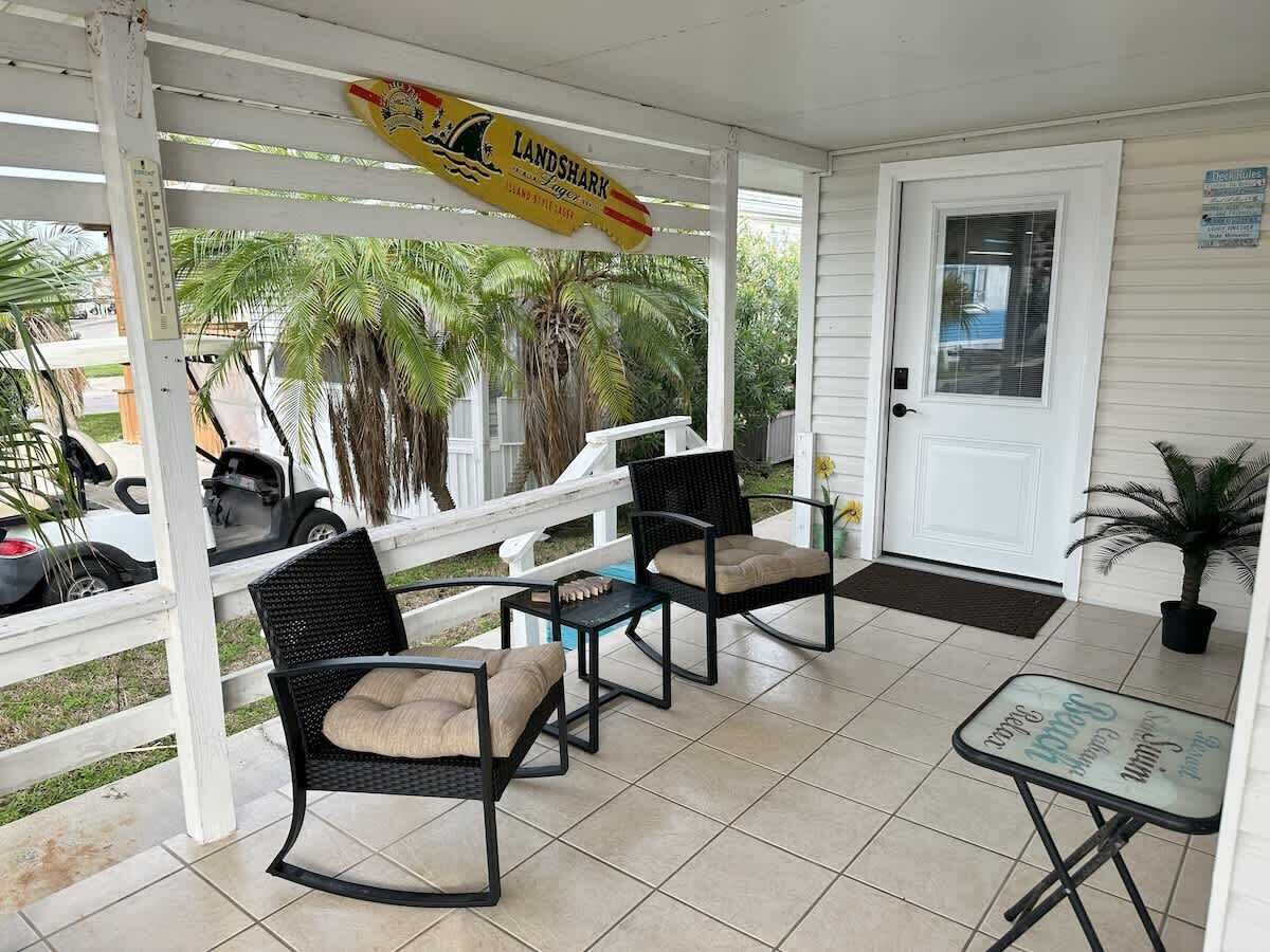 A covered porch with two wicker chairs and a small table, a white door, and a sign above.