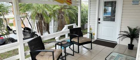 A covered porch with two wicker chairs and a small table, a white door, and a sign above.