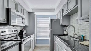 Une cuisine étroite dotée d’appareils électroménagers en acier inoxydable, de meubles blancs et d’une fenêtre avec des stores.