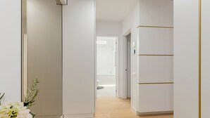 Um corredor com paredes brancas e chão de madeira, que leva a um banheiro com vaso sanitário e chuveiro.