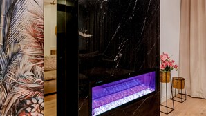 Uma sala de estar moderna com uma lareira grande de mármore preto, um papel de parede com padrão de folha tropical e piso de madeira.