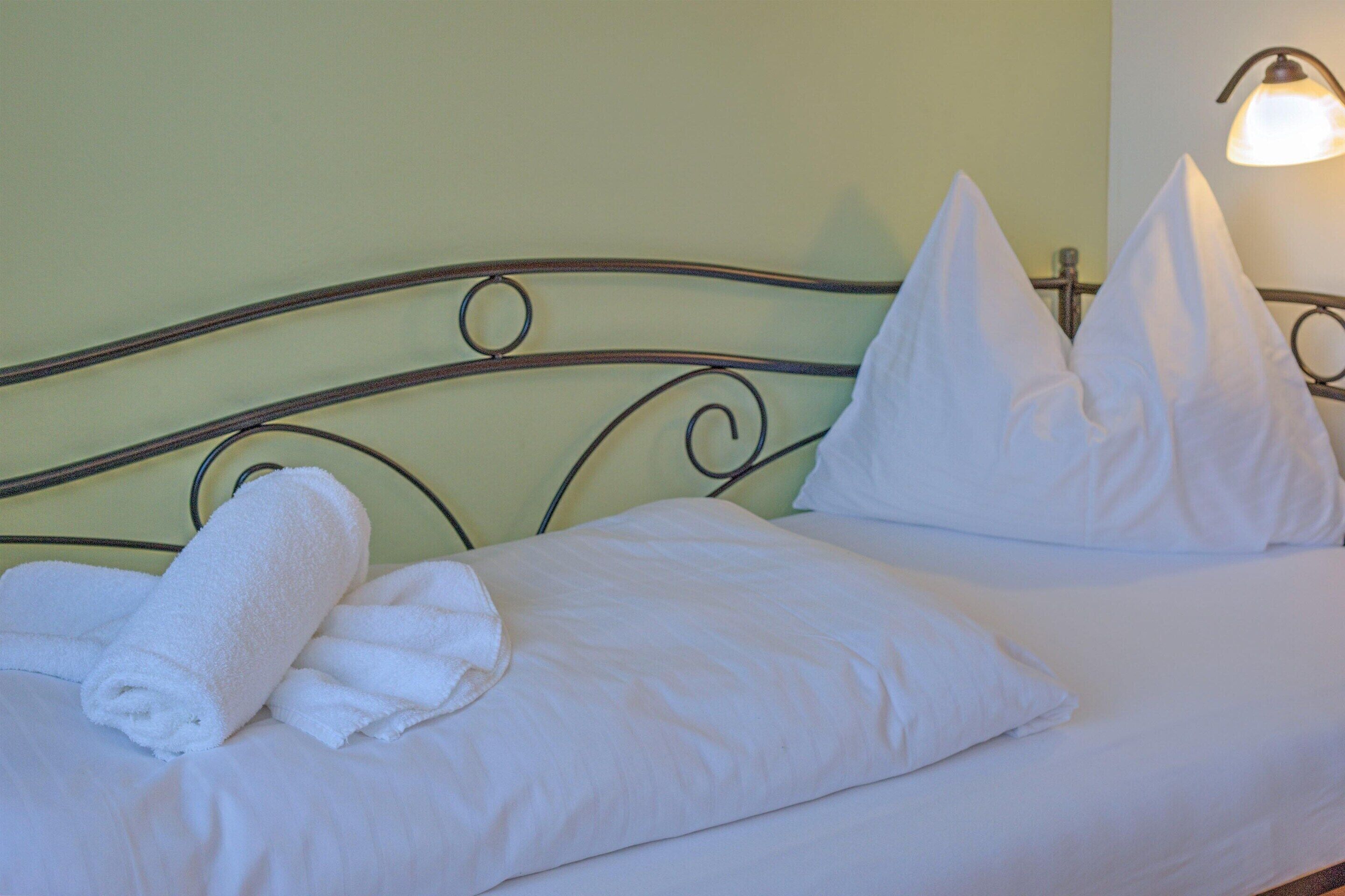 A neatly made bed with white linens and pillows, a rolled white towel, and a wall-mounted light fixture.