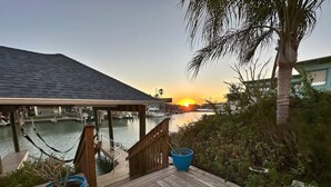 Une terrasse en bois offrant une vue sur un coucher de soleil sur l’eau, un espace abrité et deux plantes en pot.