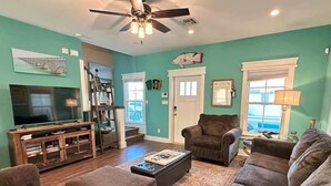 Un salon aux murs de couleur turquoise, doté d’un ventilateur de plafond, d’un téléviseur à écran plat, d’un meuble de rangement en bois et d’une grande fenêtre offrant une vue dégagée.