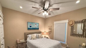 Une chambre à coucher comprenant un lit, des tables de chevet, un ventilateur de plafond, un tableau, une fenêtre avec des stores et un miroir.