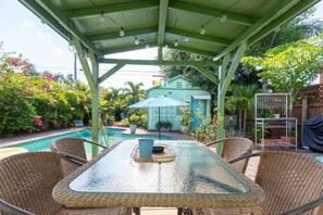 Eine überdachte Terrasse mit Glastisch und Korbstühlen, mit Blick auf einen Swimmingpool und einen üppigen Garten.
