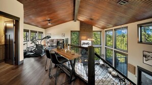 A spacious living area with a wooden ceiling, large windows offering a view of the outdoors, a dining table with chairs, and a staircase leading to an upper level. - Whispering Woods Peak.  Come see the views? (Lead)