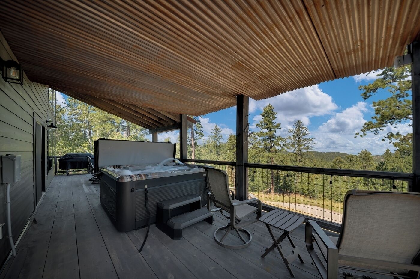 A covered outdoor deck with a hot tub, seating, and a view of trees and hills.