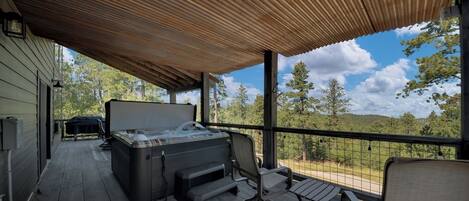 A covered outdoor deck with a hot tub, seating, and a view of trees and hills.