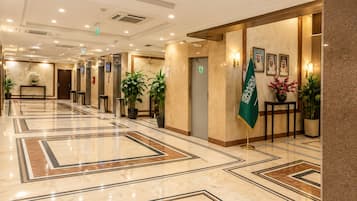 Vestíbulo de hotel con piso de mármol, ascensores, plantas en macetas y una bandera.