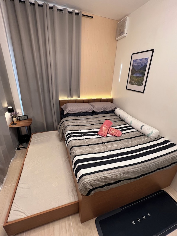 A compact hotel room with a bed, a small bedside table, a nightstand, a wall-mounted air conditioner, and a framed picture.