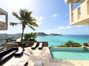 A poolside area with lounge chairs, a palm tree, and a view of the ocean and distant land. - 5BR Hilltop Villa! Pool & Ocean Views (Sint Maarten)