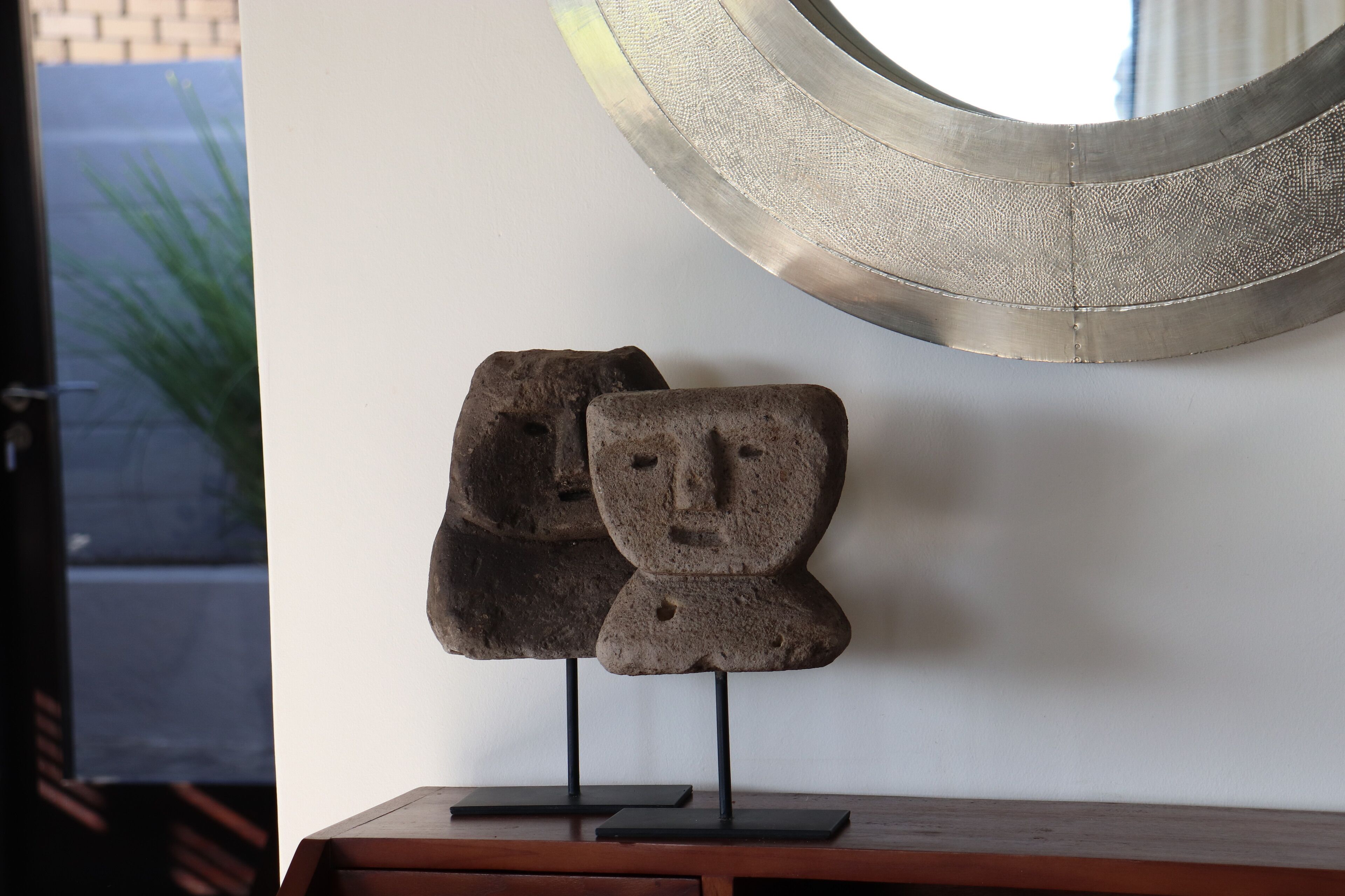Een sculptuur van twee gezichten op een houten voetstuk, tegen een muur met een ronde spiegel van metaal erboven.