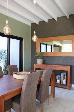 A dining area with a wooden table, grey upholstered chairs, hanging pendant lights, a large mirror, and a potted plant. - Salty Villa - Central Swakopmund Luxury 4-Bedroom Home with Private Garden (Swakopmund)