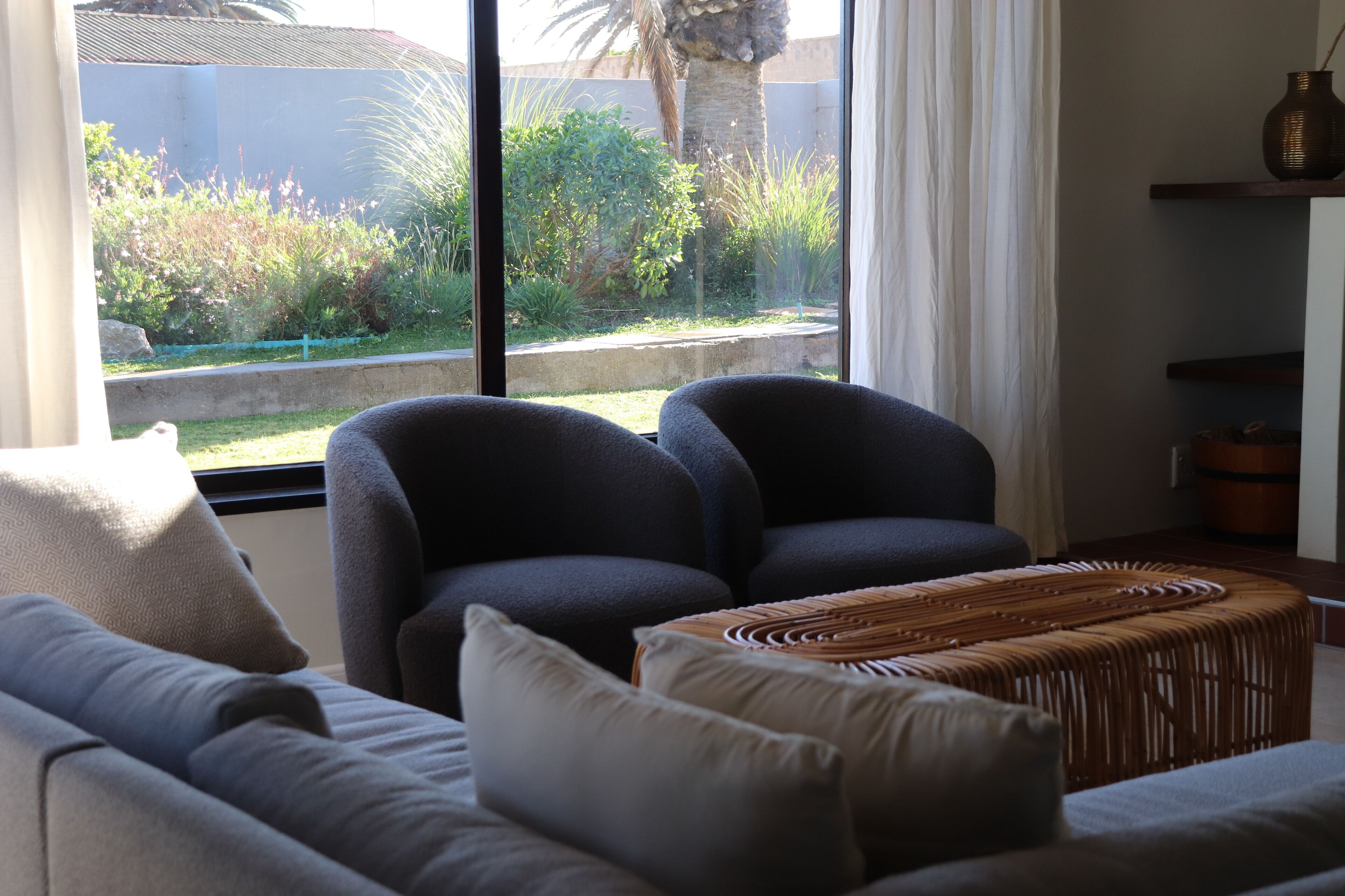 A living room with a sofa, two armchairs, a woven ottoman, and a large window with a view of greenery and a palm tree.
