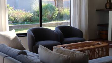 A living room with a sofa, two armchairs, a woven ottoman, and a large window with a view of greenery and a palm tree.
