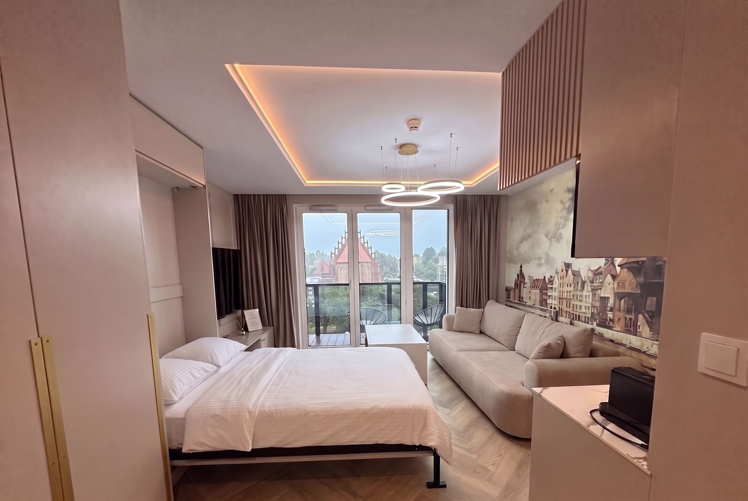 A modern hotel room with a bed, a sofa, a desk, and a view of a cityscape through large windows.