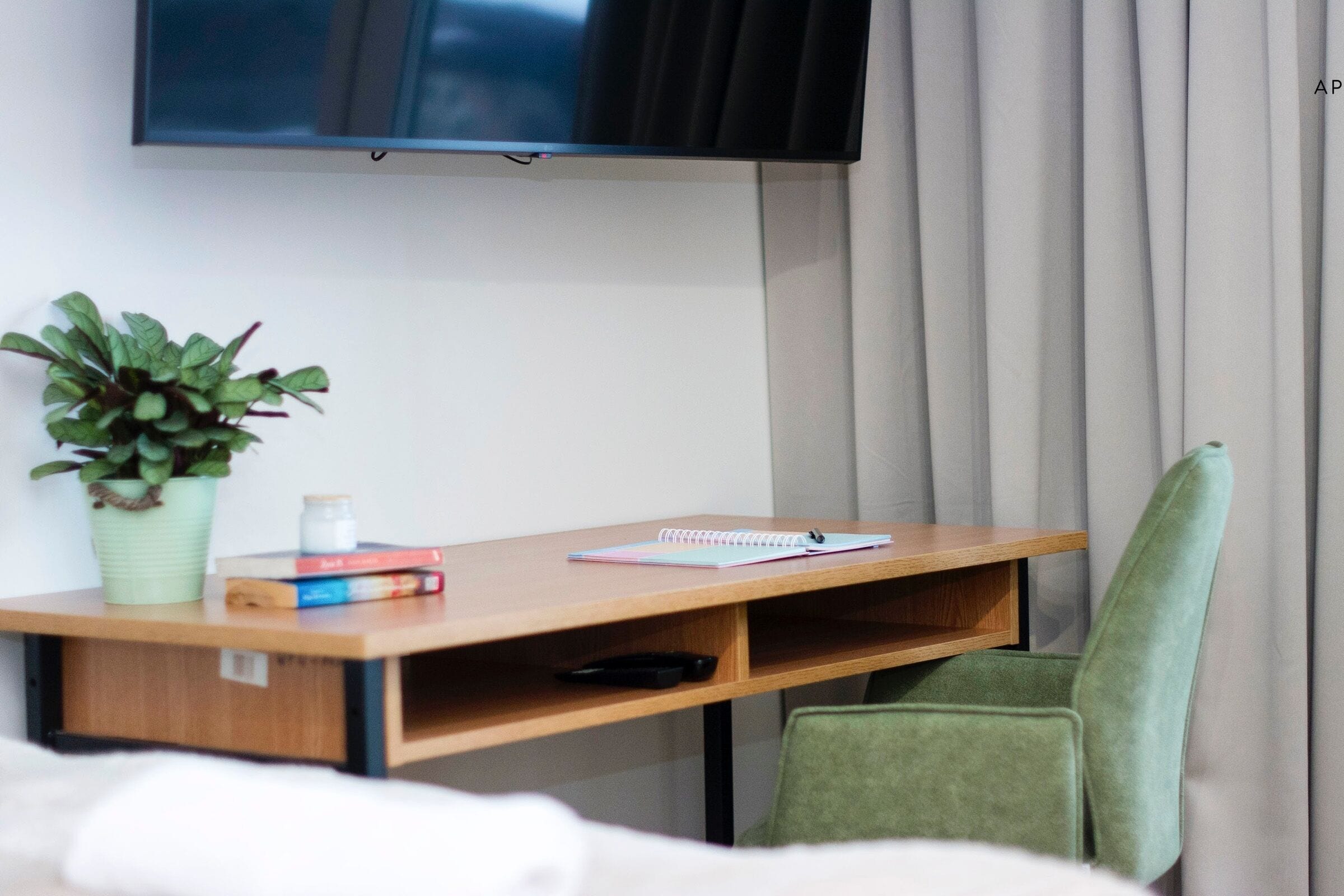 A hotel room with a wooden desk, a green chair, a potted plant, a notebook, and a television mounted on the wall.