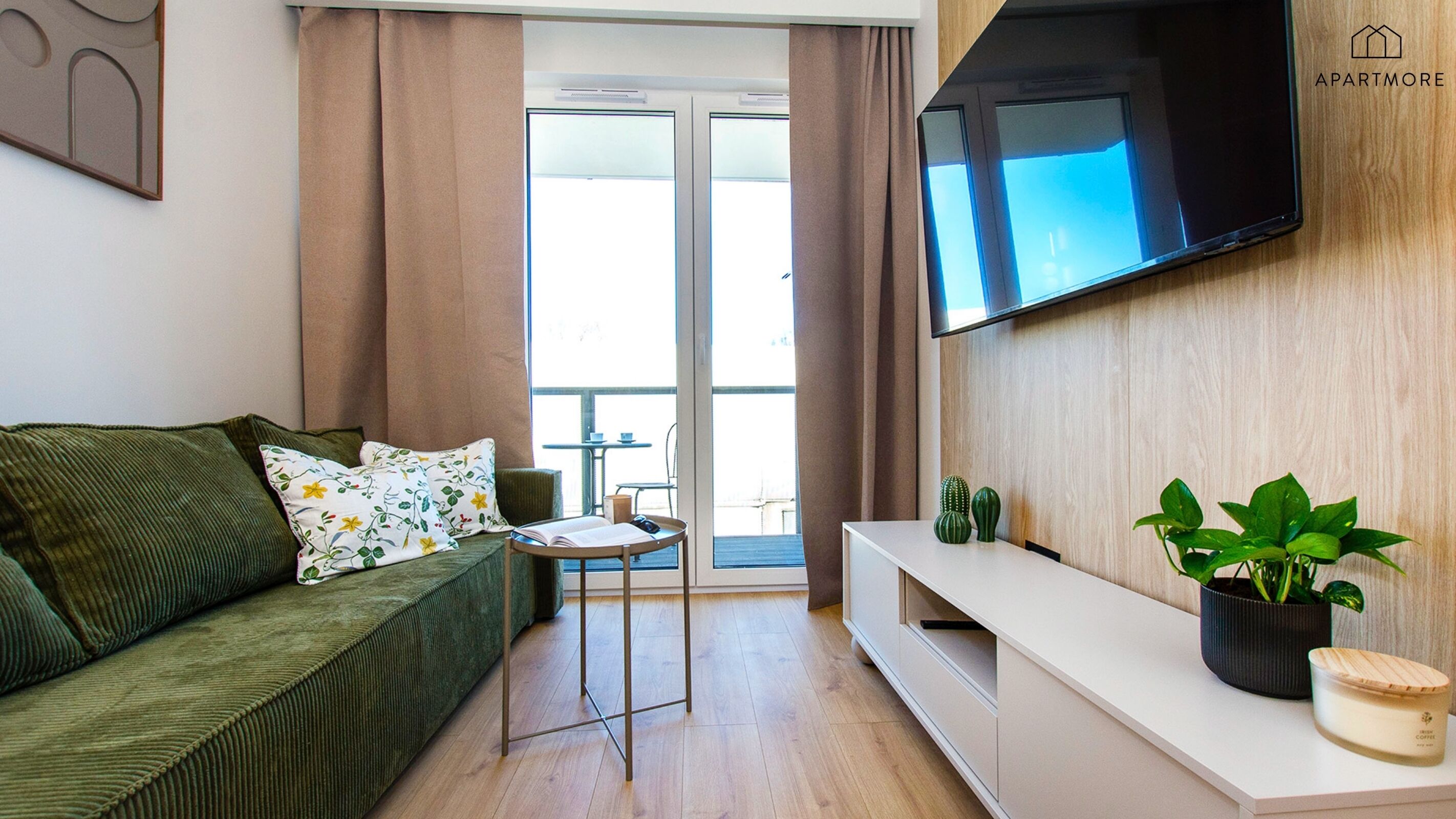 A modern living room with a green sofa, a small table, a flat-screen TV mounted on the wall, and a balcony with a view.