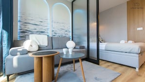 Ein modernes Schlafzimmer mit einem grauen Sofa, einem runden Holztisch und einem Bett mit aufgerollten Handtüchern.