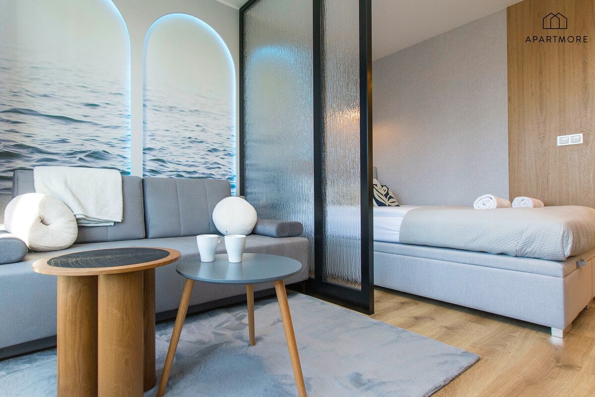 Ein modernes Schlafzimmer mit einem grauen Sofa, einem runden Holztisch und einem Bett mit aufgerollten Handtüchern.