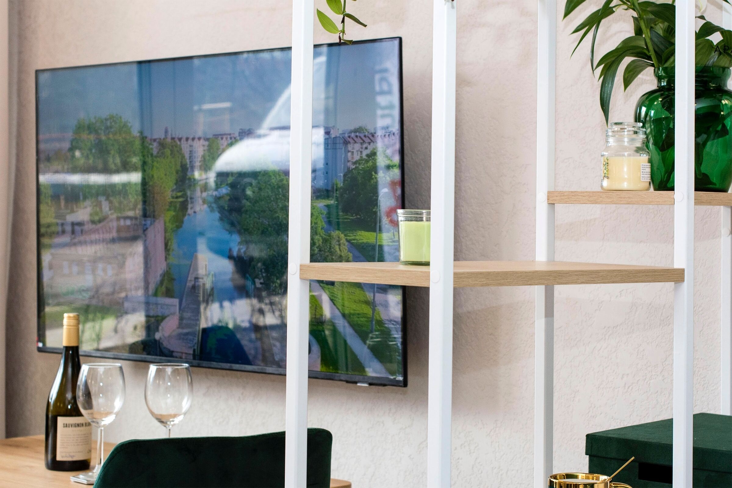 A modern room with a flat-screen TV mounted on the wall, a wooden table with a bottle of wine and glasses, a green chair, and a plant on a shelf.