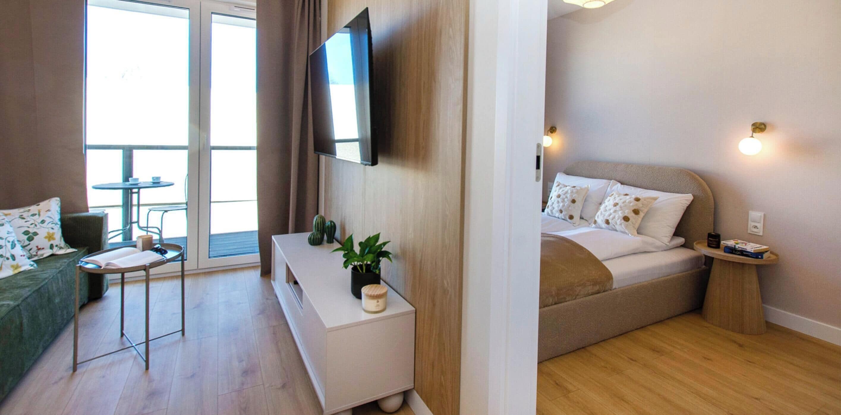 Quarto de hotel moderno com sofá, uma mesinha, mesa de cabeceira, televisão fixada na parede e cama com almofadas.