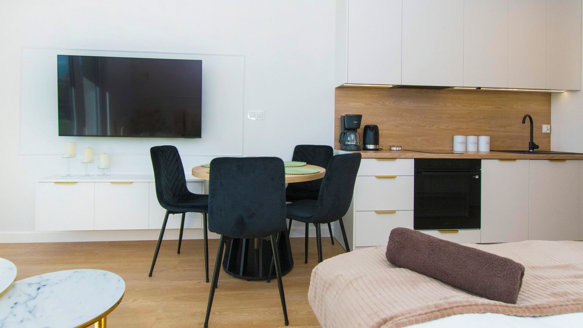 Una camera d'albergo moderna con una TV a schermo piatto, un'area pranzo con sedie, una cucina con una macchina del caffè e un letto con un cuscino.