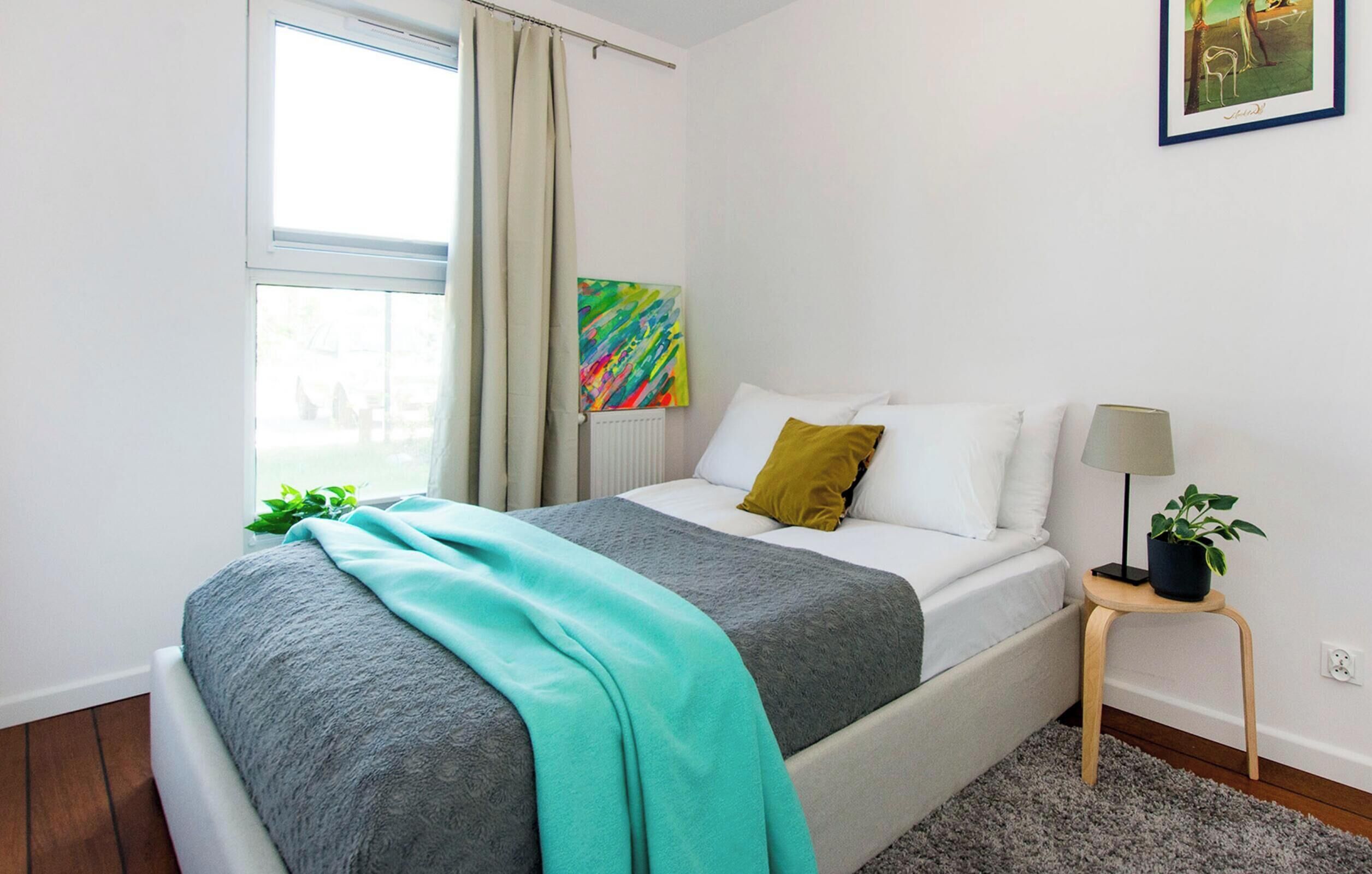 A bedroom with a bed, a nightstand, a lamp, a plant, and a painting on the wall.