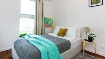 Um quarto com cama, mesa de cabeceira, abajur, planta e um quadro na parede.