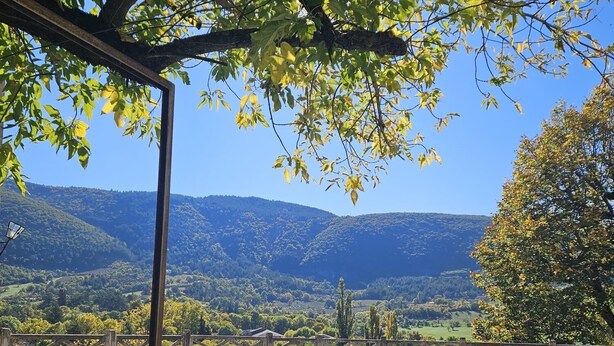 Ein Sitzbereich im Freien mit weißen Metallstühlen und -tischen, einer Pergola und Blick auf eine gebirgige Landschaft.