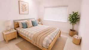 Een slaapkamer met een bed, nachtkastje, lamp, een kunstwerk in een lijst, raam met jaloezieën en potplanten.
