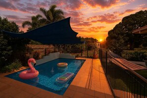 Une piscine avec vue sur le coucher de soleil, entourée d’une clôture et de palmiers.