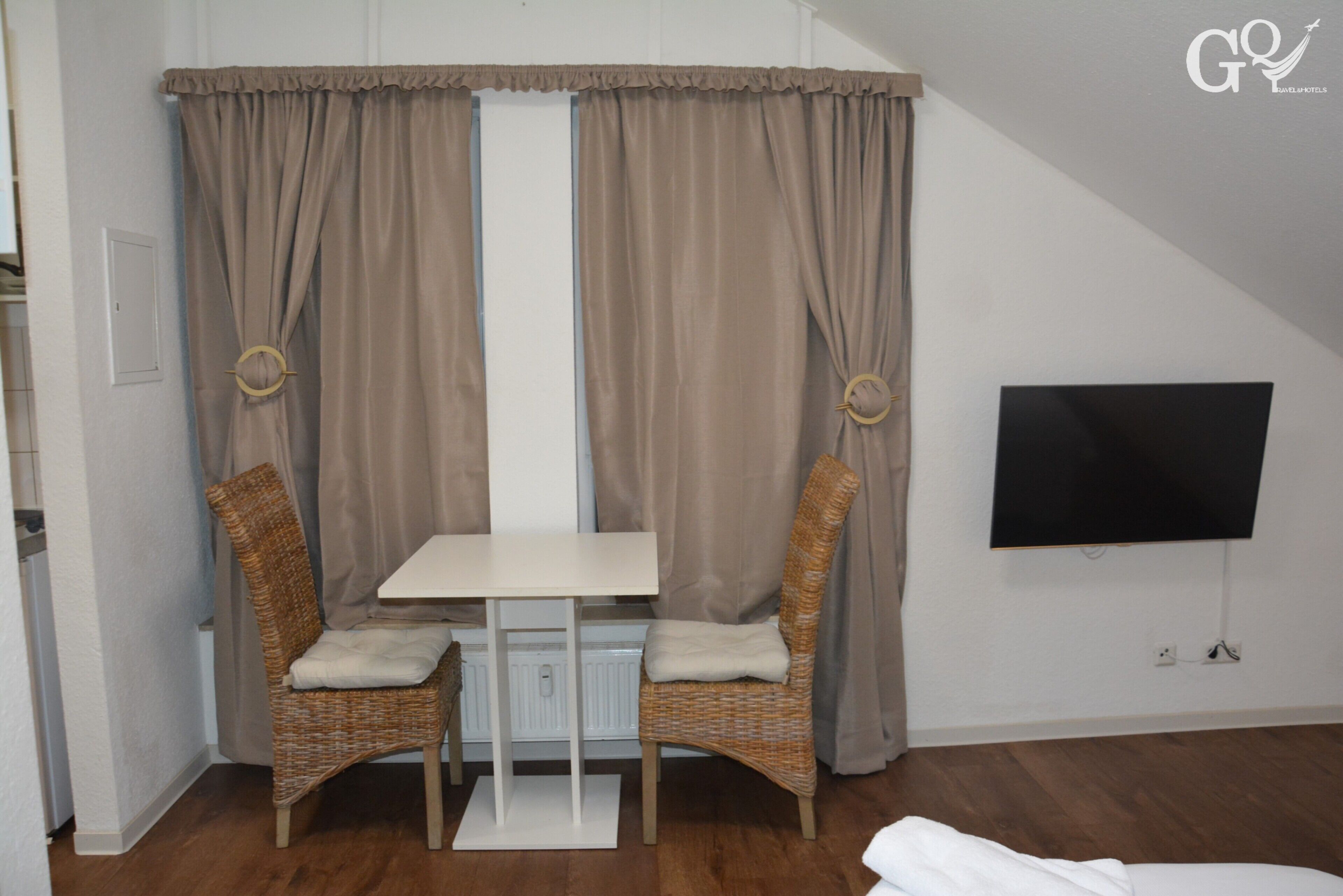Una habitació amb una taula blanca i dues cadires de vimet, un televisor fixat a la paret i una finestra amb cortines.