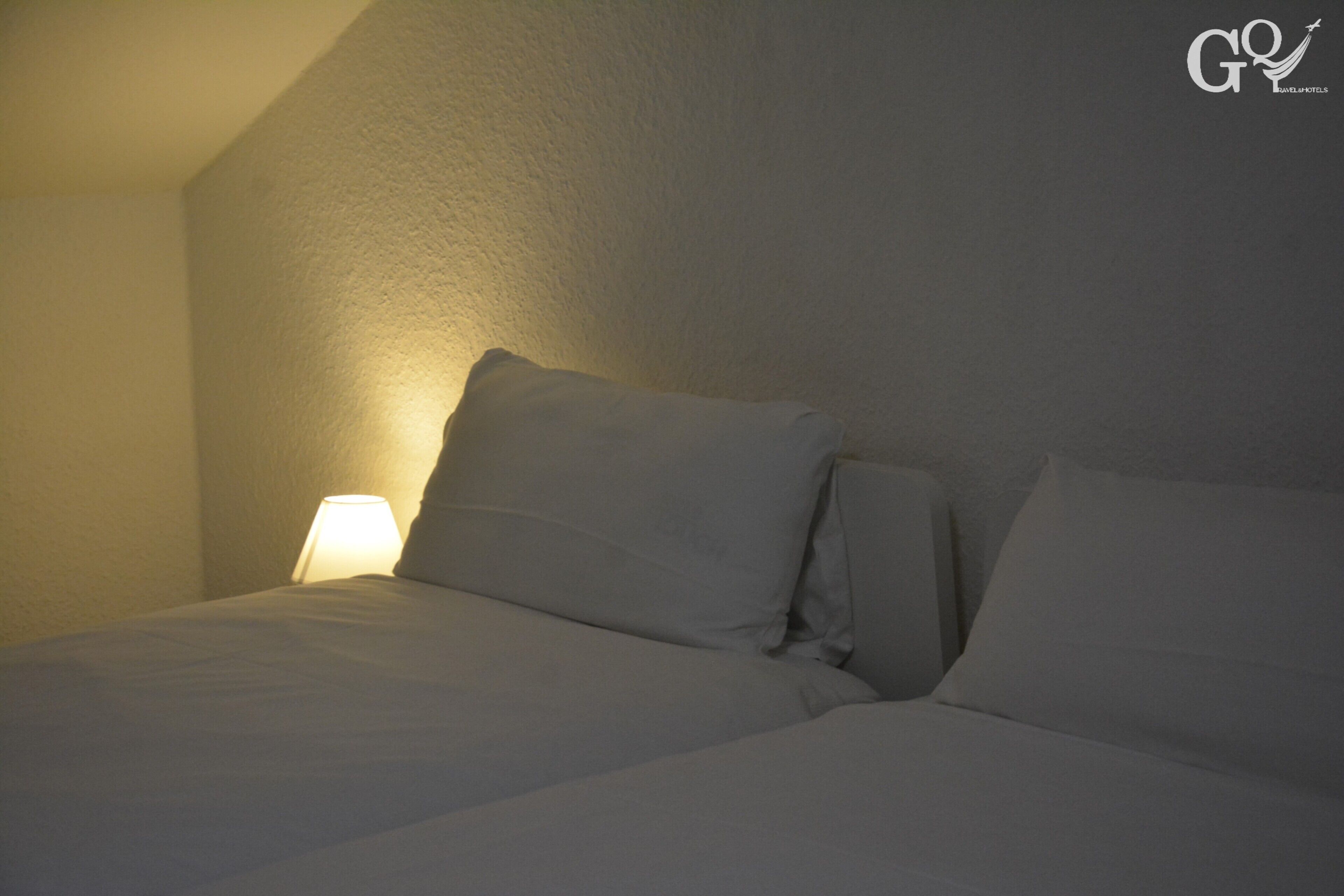 Una cama bien hecha con sábanas y almohadas blancas, una lámpara de noche y una pared lisa.