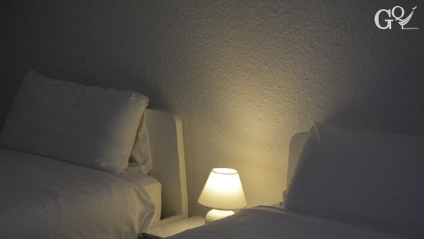 Quarto de hotel com iluminação suave, uma luminária de cabeceira, roupa de cama branca e parede com textura.